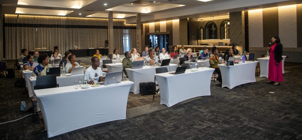 Teaching in session, teacher at the front addressing the students who are sitting at tables with white table cloths, and laptops open